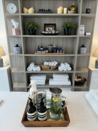 A tray on a massage table with a water pitcher with herbs and lemon.  In the background, beautifully arranged shelves with clean towels, candles and massage oils.